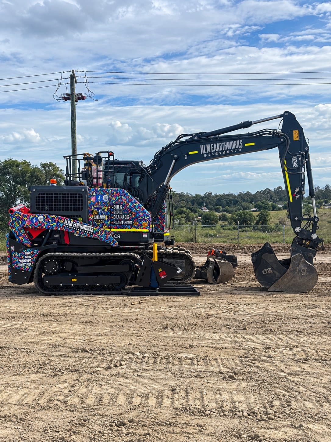 Black HLM Earthworks excavator with a brightly wrapped counterweight, sitting on a prepared dirt pad under a cloudy sky.