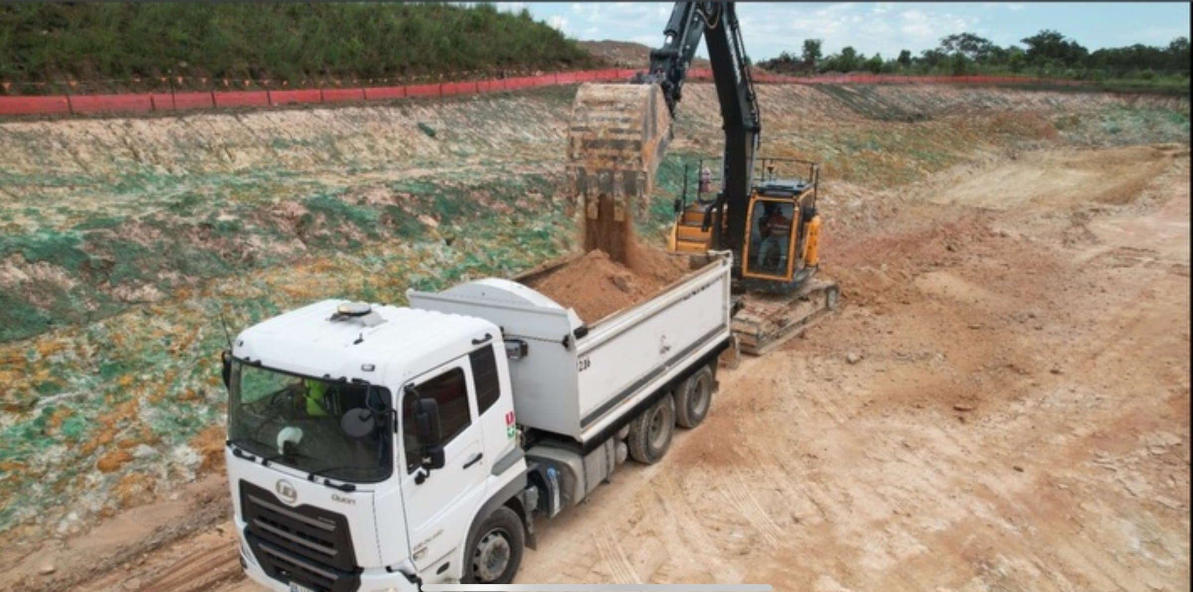 Excavator loading a tipper truck inside a deep cut, with battered faces and sediment fencing along the crest.