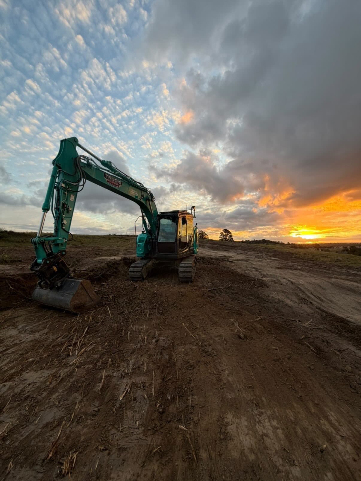 Teal Kobelco excavator working a stripped earthworks pad at sunset, with storm light breaking on the horizon.