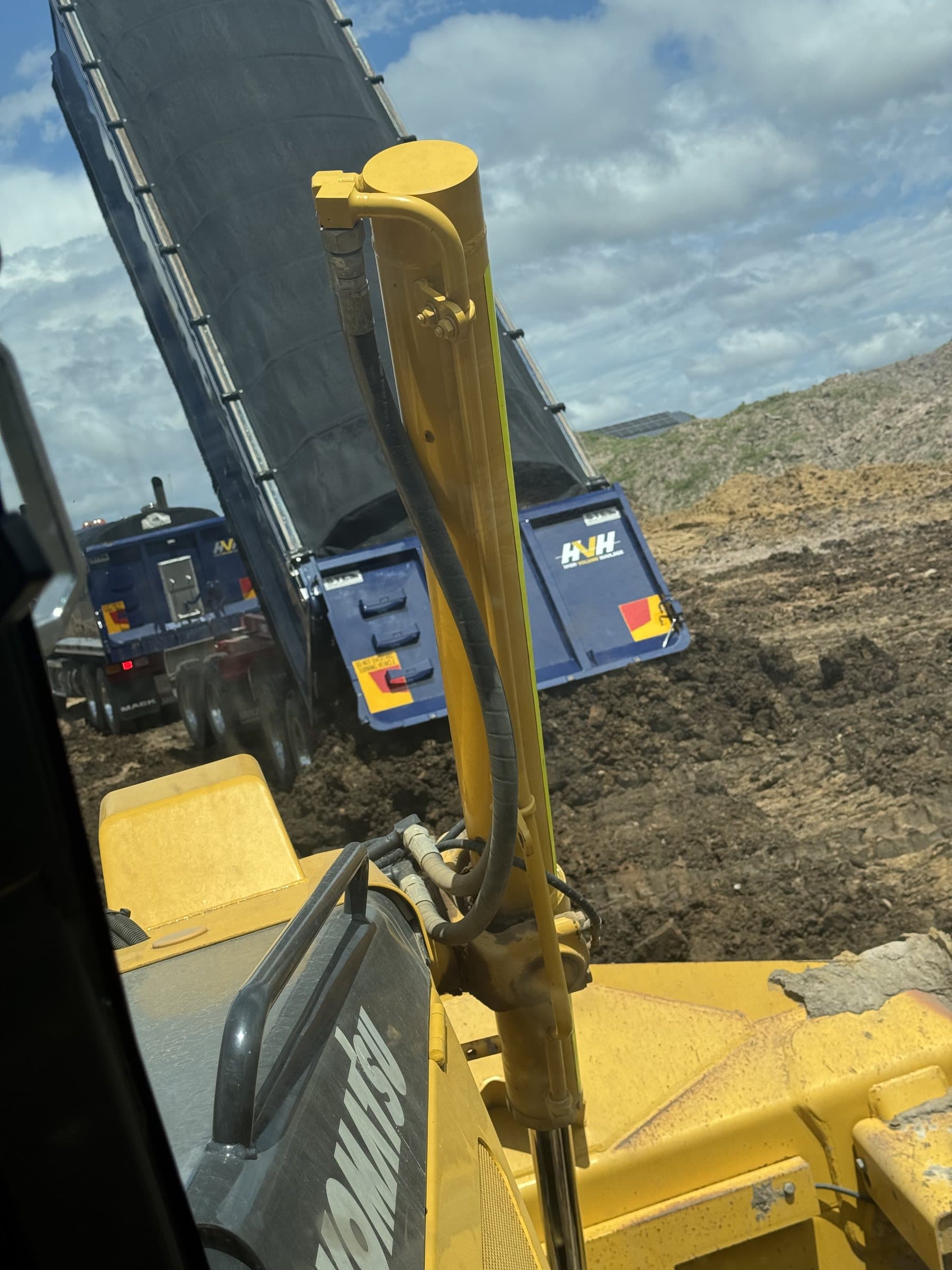 View from a Komatsu dozer cab as a blue truck-and-dog tips fill on a bulk earthworks site.